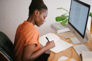 Woman Working Remotely Writing in Notebook in Front of a Computer